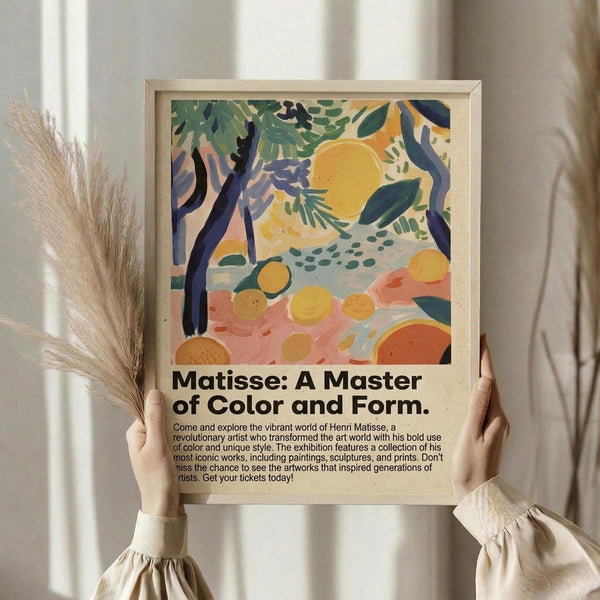 May include: Person holding framed art print featuring abstract landscape with yellow sun, green foliage, orange fruits, and purple tree branches in Matisse-inspired style. Light wood frame against neutral curtained background with dried pampas grass visible. Cream-colored sweater with gathered sleeves.