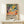 May include: Framed Matisse exhibition poster featuring tropical landscape with orange sun, green foliage, dark tree trunks, and scattered oranges on pink ground. Poster displays in thin wooden frame on light wood console table beside small white gift box with greenery sprig against cream wall.