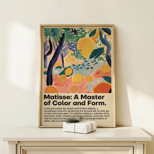 May include: Framed Matisse exhibition poster featuring tropical landscape with orange sun, green foliage, dark tree trunks, and scattered oranges on pink ground. Poster displays in thin wooden frame on light wood console table beside small white gift box with greenery sprig against cream wall.
