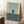 May include: Framed abstract art print leaning against white wall in cozy living room setting. Artwork features geometric composition with pale green background, dark blue rectangle, and yellow border accents. Natural wood frame complements minimalist decor with coffee cup on wooden side table and textured cream rug below.