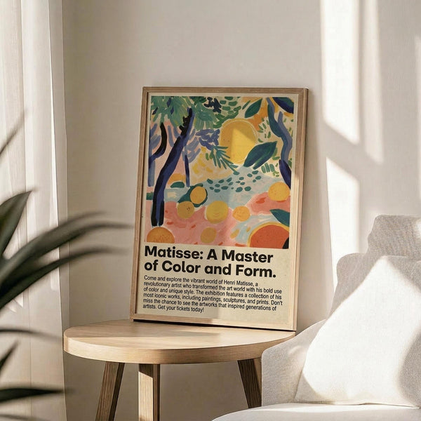 May include: Colorful abstract art poster in wooden frame displaying tropical foliage, fruits, and organic shapes in yellow, green, pink, and blue tones. Frame sits on round wooden side table next to beige upholstered chair with plant leaves visible in corner. Natural sunlight creates soft shadows on neutral wall background.