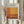May include: Person holding framed exhibition poster for Mark Rothko featuring abstract orange and rust colored rectangular color fields. Light wood frame against neutral curtained background with dried pampas grass stems visible on left side. Warm earth tones throughout composition.