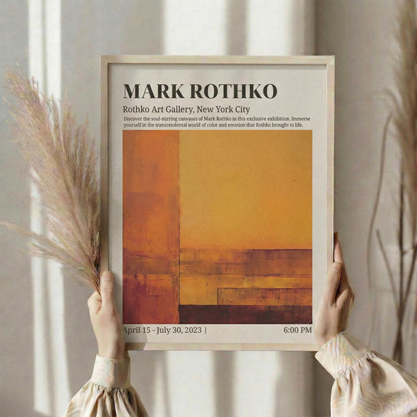 May include: Person holding framed exhibition poster for Mark Rothko featuring abstract orange and rust colored rectangular color fields. Light wood frame against neutral curtained background with dried pampas grass stems visible on left side. Warm earth tones throughout composition.