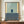 May include: Framed abstract art print displayed on wooden sideboard in elegant room with white paneled walls. The minimalist artwork features geometric blocks of muted blue-green and darker blue against cream and yellow sections. A small white ceramic vase and stack of books sit nearby on the warm wood surface.
