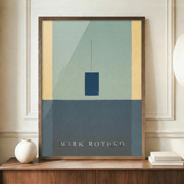 May include: Framed abstract art print displayed on wooden sideboard in elegant room with white paneled walls. The minimalist artwork features geometric blocks of muted blue-green and darker blue against cream and yellow sections. A small white ceramic vase and stack of books sit nearby on the warm wood surface.