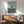 a living room with a wooden cabinet, a green armchair, and a wooden coffee table. Above the cabinet, there is a framed abstract art piece with a blue and green color scheme.