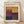May include: Framed exhibition poster featuring Mark Rothko's name in bold black letters at top, with colorful abstract color block artwork below in warm yellows, deep reds, purples, and dark blues arranged in rectangular segments. Poster shows gallery details and exhibition dates. Light wood frame sits on wooden floor against white wall with soft natural lighting and shadows.
