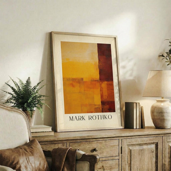 May include: Framed abstract art print with orange, yellow, and burgundy color blocks in warm earth tones, displayed on rustic wooden sideboard alongside fern plant, ceramic vase, and stacked books in cozy living room with neutral beige chair and soft natural lighting.