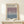 May include: A framed exhibition poster for Mark Rothko displayed on a light wood shelf against a cream wall. The poster features a gradient design transitioning from purple at top to blue at bottom, with black text and exhibition details. The thin wood frame complements the minimalist aesthetic. A small white wrapped gift with natural twine and greenery sits beside the frame on the shelf.