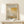 May include: Framed exhibition poster with Mark Rothko's name in bold black text, featuring abstract geometric artwork in warm yellows, oranges, and muted browns. Light wood frame displayed on white wooden surface against cream wall, with small wrapped gift box beside it. Natural lighting creates soft shadows.