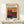 May include: Framed exhibition poster featuring Mark Rothko's abstract color field artwork with geometric blocks in warm yellows, deep reds, purples, and dark blues. Light wood frame displayed on white floating shelf against cream wall with small wrapped gift box beside it. Soft natural lighting creates gentle shadows.