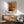 May include: Framed abstract art poster with warm orange, yellow, and amber color blocks in Rothko's signature style, displayed above a wooden sideboard with white ceramic lamp. Mid-century modern living room setting features sage green upholstered chair with wooden arms and round coffee table. Soft natural lighting creates shadows across white walls.