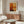 May include: Abstract art poster with warm orange, yellow and deep red rectangular color blocks in minimalist wooden frame, displayed above mid-century modern walnut credenza in neutral living room with white curved chair, glass coffee table, and modern brass floor lamp.