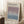 May include: Framed poster leaning against white wall on fluffy white rug. Features gradient artwork transitioning from purple at top to blue at bottom. Text reads "MARK ROTHKO" and "Rothko Art Gallery, New York City" with exhibition dates "April 15 - July 30, 2023" and "6:00 PM". Light wood frame, cozy room setting with coffee cup on wooden block and beige blanket visible.