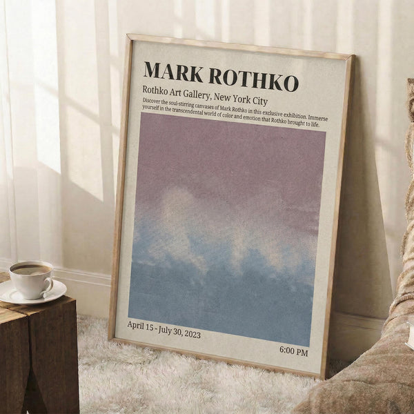 May include: Framed poster leaning against white wall on fluffy white rug. Features gradient artwork transitioning from purple at top to blue at bottom. Text reads "MARK ROTHKO" and "Rothko Art Gallery, New York City" with exhibition dates "April 15 - July 30, 2023" and "6:00 PM". Light wood frame, cozy room setting with coffee cup on wooden block and beige blanket visible.