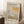 May include: Framed Mark Rothko exhibition poster leaning against white wall corner on fluffy cream rug. Poster features abstract rectangular blocks in warm yellow, beige and gray tones with black text header and exhibition details. Light wood frame, cozy room setting with coffee cup on wooden block and beige textured throw blanket visible.