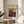 May include: A framed exhibition poster for Mark Rothko's art gallery show displayed on a round wooden side table next to a beige upholstered chair. The poster features abstract color blocks in warm yellows, deep reds, purples, and navy blue arranged in rectangular sections. Sunlight streams across the neutral-toned room, casting shadows from a plant's leaves. The wooden frame matches the natural wood table, creating a cohesive modern interior design aesthetic.