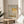 May include: Framed exhibition poster featuring Mark Rothko's abstract artwork in soft yellow and beige rectangular color blocks, displayed on a round wooden side table next to a beige upholstered chair with white pillow. Natural sunlight streams across the neutral-toned room setting with plant leaves visible in the corner.