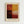 May include: Framed abstract art poster with six color blocks in warm earth tones - deep reds, golden yellow, dark burgundy, cream, and muted orange sections with painterly brushstroke textures. Artist name printed below artwork. Light wood frame against white wall in modern interior setting.