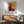 May include: Framed Mark Rothko abstract art print featuring blocks of warm red, orange, yellow, and burgundy colors hanging above a mid-century modern wooden credenza with white ceramic lamp. Light blue upholstered armchair with wooden frame sits nearby on wooden floors with natural sunlight casting shadows across white walls.