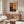 May include: Abstract art poster with red, yellow, orange and dark brown color blocks in grid pattern, displayed above mid-century modern wooden credenza in minimalist living room. White curved chair and round glass coffee table visible. Warm natural lighting creates shadows on light gray walls.