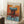 May include: Abstract expressionist poster in light wood frame leaning against white wall corner. Artwork features vibrant orange background with turquoise blue horizontal brushstroke and dark red-brown vertical form below, creating window-like shape. Blue paint drips visible at bottom. White coffee cup on wooden block nearby, cream textured rug and beige fabric visible. Warm natural lighting creates soft shadows on white walls.