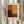 May include: Person in beige ruffled sleeve top holding framed abstract art print featuring six color blocks in warm tones - red-orange, golden yellow, dark burgundy, cream, coral red, and deep green arranged in a grid pattern. Light wooden frame with cream matting. Dried pampas grass visible on left side against soft neutral curtained background.