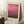 May include: Hands holding a framed art print featuring a pink and burgundy gradient color field painting in minimalist style. The print has "MARK ROTHKO" text at bottom and is displayed in a cozy room setting with white walls, wooden floor, cream rug, wooden side table with coffee cup, and beige throw blanket visible.