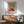 May include: Abstract art poster with orange and teal geometric blocks in warm wood frame, displayed above mid-century modern credenza with ceramic lamp, next to mint green upholstered chair with wooden arms in bright sunlit room with white walls