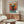 May include: Abstract expressionist poster with vibrant orange background and blue-green rectangular forms arranged vertically, displayed above a mid-century modern walnut credenza. Room features cream textured accent chair, glass oval coffee table, sculptural floor lamp, and table lamp with white shade. Bright natural lighting creates shadows across polished floors.