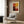 May include: Abstract color block artwork in warm reds, oranges, and yellows displayed in cream matted frame on wood paneled wall above beige upholstered armchair with geometric patterned pillow, brass floor lamp, and dark round side table with open book in modern living room setting with natural lighting from window.