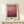 May include: Abstract gradient artwork in warm wood frame leaning against cream wall on light wooden surface. Image shows coral pink at top transitioning through magenta to deep burgundy and brown at bottom with horizontal brushstroke texture. Small white wrapped gift with green sprig sits beside frame. Soft natural lighting creates gentle shadows.