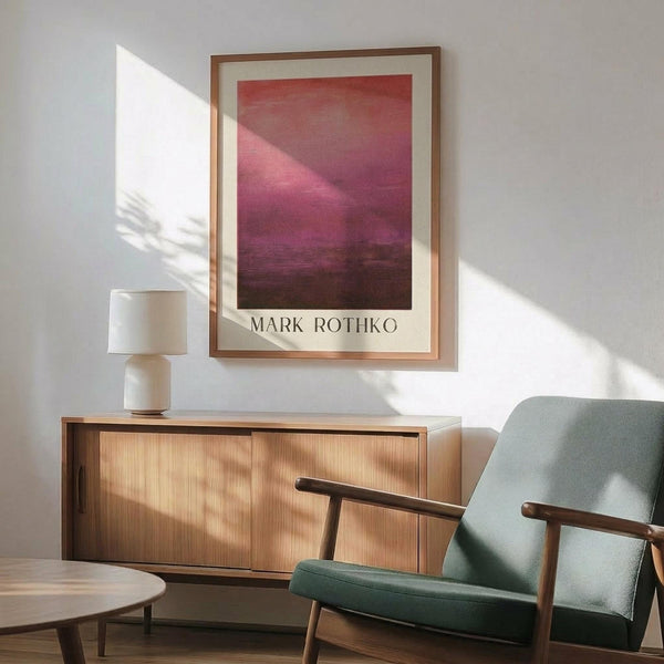 May include: Framed Mark Rothko poster with pink and burgundy abstract color fields hanging above modern wooden credenza. Mid-century sage green upholstered armchair with wooden frame sits nearby. White ceramic table lamp and round coffee table visible. Warm natural lighting creates geometric shadows on white wall.