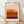 May include: Framed art print leaning against white textured wall on light wood floor. Print features horizontal color blocks in warm earth tones - light blue sky at top transitioning through peachy beige, golden yellow, burnt orange with textured brushwork, and deep burgundy at bottom. Natural wood frame with white matting. Soft natural lighting creates diagonal shadows across the scene.