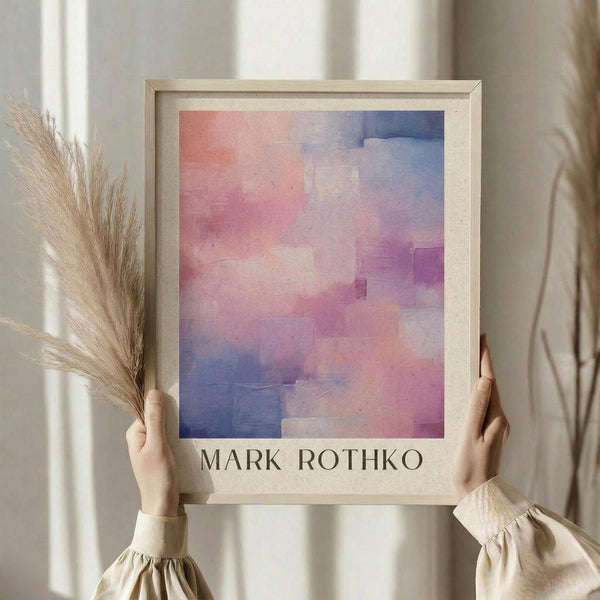 May include: Person holding framed abstract art print with soft gradient colors blending from coral pink to deep blue and purple in pixelated blocks. Light wood frame with cream matting displays artist name below artwork. Dried pampas grass stems held in other hand against neutral beige curtained background.