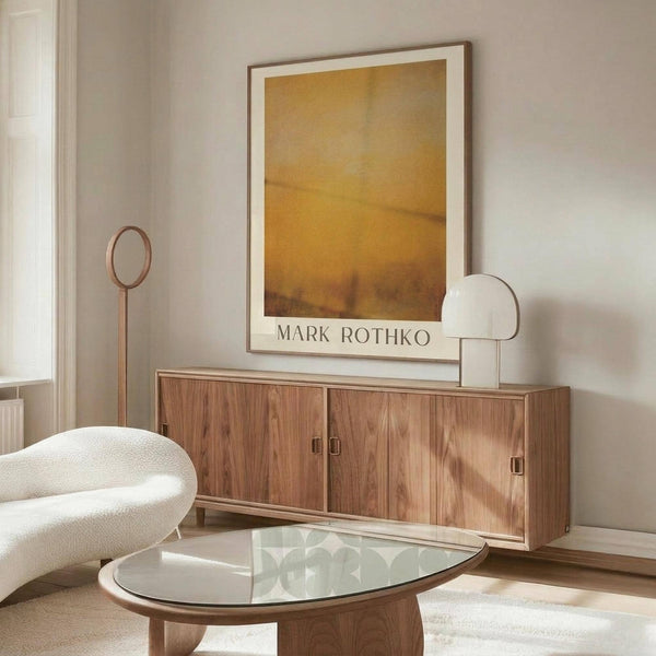 May include: A framed Mark Rothko poster print with warm golden yellow and amber tones displayed above a mid-century modern walnut credenza. The room features minimalist decor with a cream upholstered chair, glass-top coffee table, floor lamp, and table lamp. Soft natural lighting creates shadows on the neutral walls, showcasing the artwork's rich sunset-like colors in a contemporary living space.