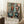 May include: Framed abstract art poster leaning against white wall corner on fluffy white rug. Artwork features geometric shapes in teal, orange, black and white with loose brushwork style. Coffee cup on wooden stool nearby, beige throw blanket visible. Natural lighting creates soft shadows on walls.