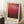 May include: Framed Mark Rothko abstract art print leaning against white wall corner in cozy room setting. Red and burgundy color field painting with gradient from bright red at top to deep maroon at bottom. Light wood frame with cream matting. White coffee cup on wooden block nearby, beige knit throw blanket visible. Soft natural lighting creates gentle shadows on textured white rug.