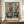 May include: Abstract modernist artwork poster in wooden frame displaying geometric kitchen objects - coffee pots, cups, and bowls in muted teal, coral, and cream tones with bold black outlines. The framed print sits on a warm wood sideboard alongside a small white ceramic vase and stacked books in a bright room with white paneled walls.
