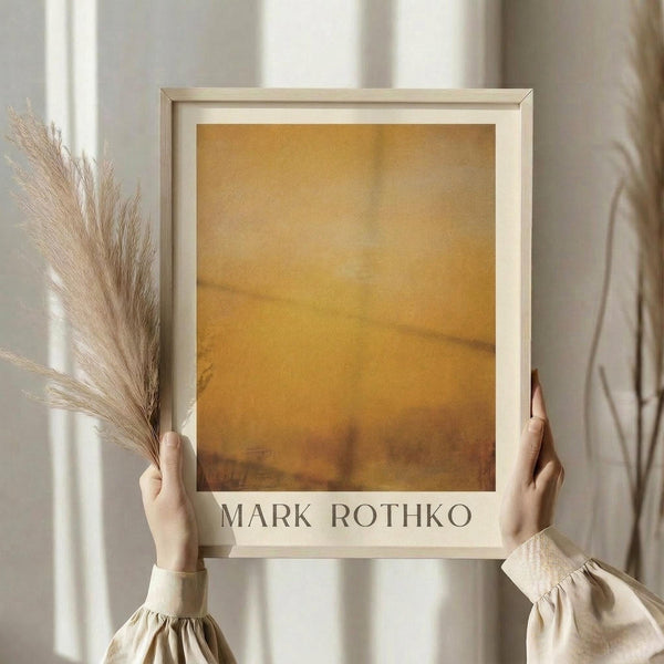 May include: Hands holding framed abstract art print with warm golden yellow and amber tones in minimalist style. Light wood frame against neutral beige curtain backdrop. Person wearing cream colored sweater with gathered cuffs also holds dried wheat stalks, creating soft natural styling scene.