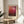 May include: Framed art poster with red and burgundy gradient abstract painting displayed on round wooden side table next to beige upholstered chair. Sunlight streams across white wall creating geometric shadows. Green plant leaves visible in corner. Poster shows deep red tones transitioning from bright coral to dark maroon with artist name below image.