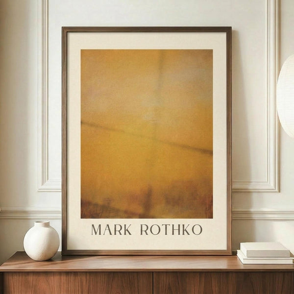 May include: Framed abstract art print in warm golden and amber tones with soft, blended brushstrokes displayed on wooden sideboard. White ceramic vase and books positioned nearby. Cream walls with decorative molding visible in background. Print features artist name text below artwork in clean typography.