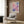 May include: Abstract watercolor artwork in pink, purple and blue tones displayed in light wood frame on wood paneled wall. Modern beige armchair with geometric patterned pillow sits below artwork next to brass floor lamp and dark round side table with open book.