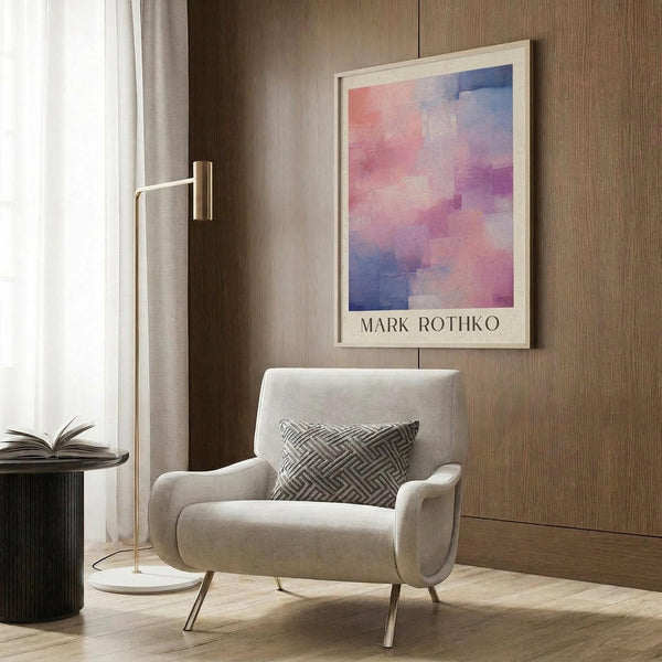May include: Abstract watercolor artwork in pink, purple and blue tones displayed in light wood frame on wood paneled wall. Modern beige armchair with geometric patterned pillow sits below artwork next to brass floor lamp and dark round side table with open book.