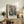 May include: Framed abstract art print featuring geometric vessels and containers in muted teal, orange, and brown tones, displayed on rustic wooden console alongside fern plant, ceramic vase, and books in warm-lit living room with cream upholstered furniture and neutral walls.
