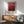 May include: Abstract red and burgundy artwork with Mark Rothko's name displayed below, hanging in wooden frame on white wall above mid-century modern wooden credenza. Green upholstered armchair with wooden arms sits nearby with white ceramic table lamp and round wooden side table. Natural sunlight creates geometric shadows across the minimalist living room scene.