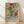 May include: Framed poster with colorful abstract floral design in Matisse-inspired style, featuring vibrant red, yellow, teal, and purple botanical shapes on cream background. Black text below reads "Celebrating Colors" with descriptive paragraph. Natural wood frame sits on light wooden floor against white wall with soft shadows.