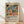 May include: Framed colorful abstract art print leaning against white wall on wooden floor. Artwork features vibrant botanical shapes including tropical leaves, fruits, and flowers in blues, greens, oranges, and reds with Matisse-inspired cut-out style. Light wood frame with cream matting. Text below reads "Celebrating Colors of Life" with product description. Soft natural lighting creates diagonal shadows on wall.