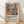 May include: Framed art print leaning against white wall on light wood floor. The poster features a Matisse-inspired illustration of a woman in coral dress sitting among potted plants and flowers in bold, flat colors including coral, navy, green, and cream. Below the artwork is black text reading "Celebrating Colors of Life" with descriptive paragraph. Natural wood frame with beige matting. Soft window light creates diagonal shadows across the white textured wall.