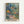 May include: Framed art print displayed on white wall featuring vibrant tropical garden scene with overlapping translucent layers in blues, greens, oranges, and corals. Abstract botanical elements include palm fronds, flowering plants, and small human figures on a winding path. Artist name "MATISSE" appears in bold letters at bottom. Natural wood frame surrounds the colorful layered composition with dreamy, collage-like aesthetic.
